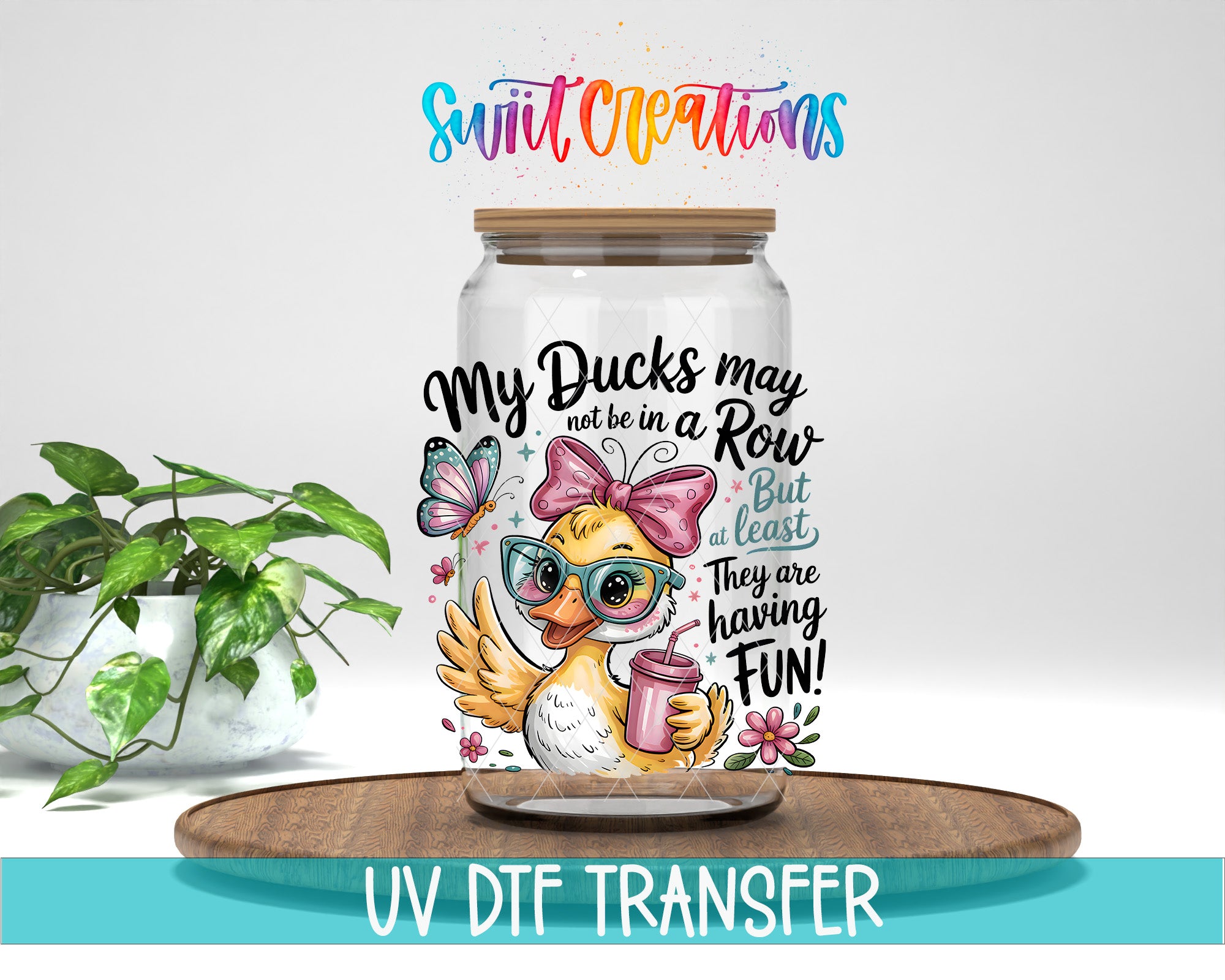 A glass jar with a cartoon duck wearing sunglasses and holding a drink, surrounded by various decorative elements such as flowers and butterflies. The text on the jar reads "My Ducks may not be in a Row but at least they are having fun!" and "Swift Creations". The jar is placed on a wooden tray, and there is a plant in the background.