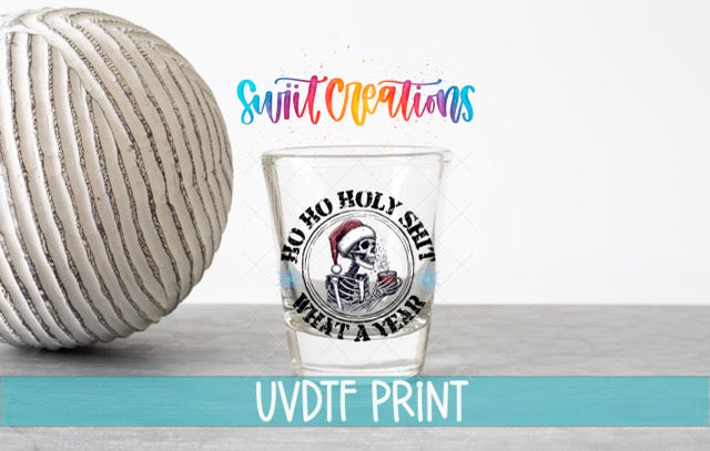 a shot glass with a skull and crossbones design, along with the text "Hoo ho holy shit what a year" printed on it. The shot glass is placed next to a ball, and there is text in the background that reads "Swit Creations" and "UVDF PRINT".