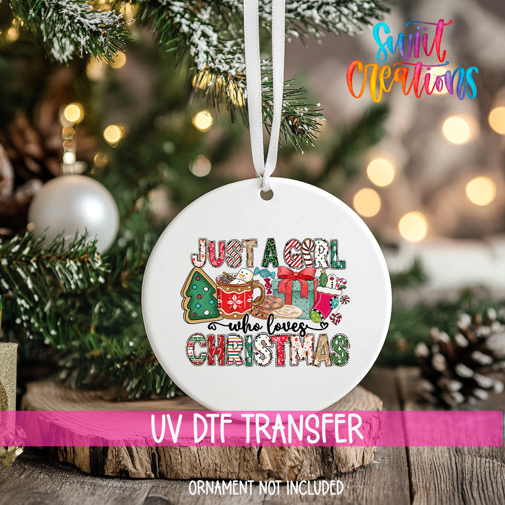 A white ceramic ornament with a colorful Christmas-themed design hangs on a tree branch, with a blurred background of lights and greenery.