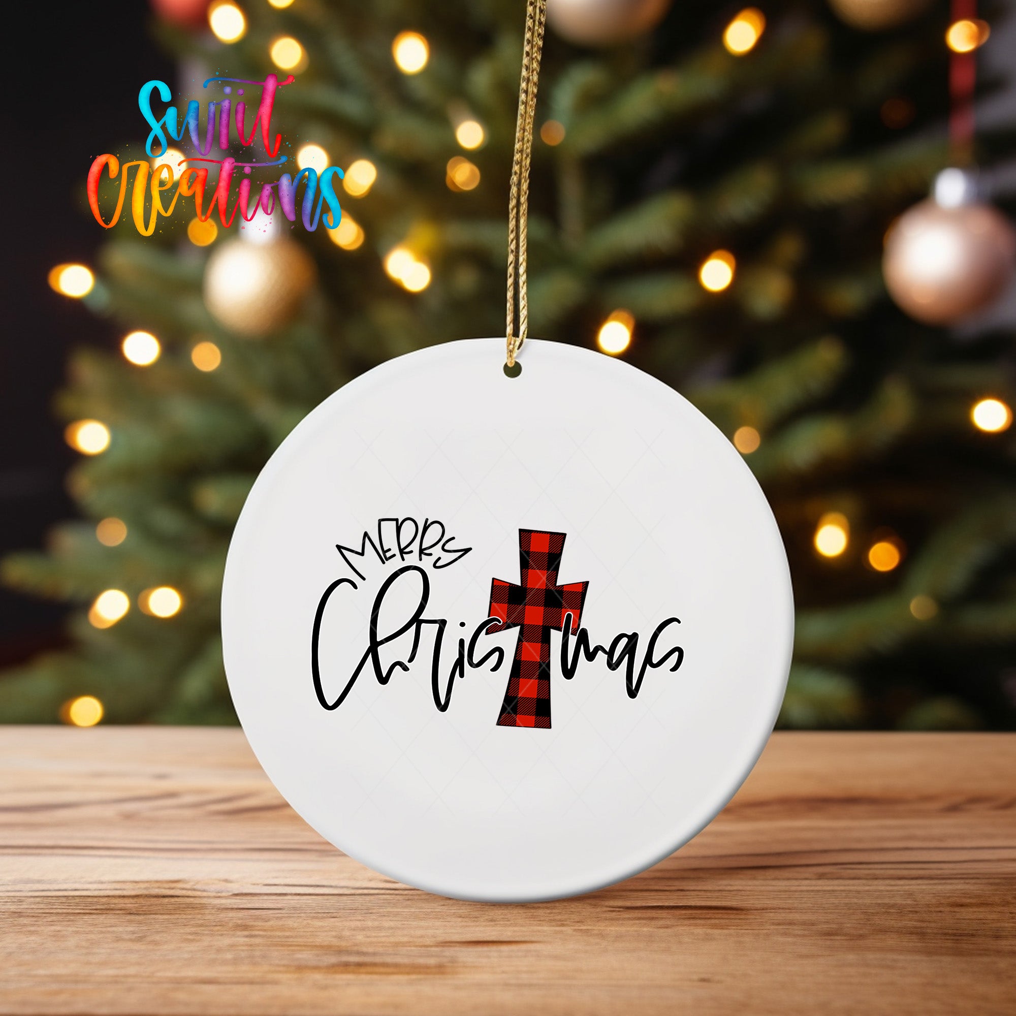 A white Christmas ornament with the text "Merry Christmas" and a red and black plaid cross design hangs in front of a blurred Christmas tree with twinkling lights.