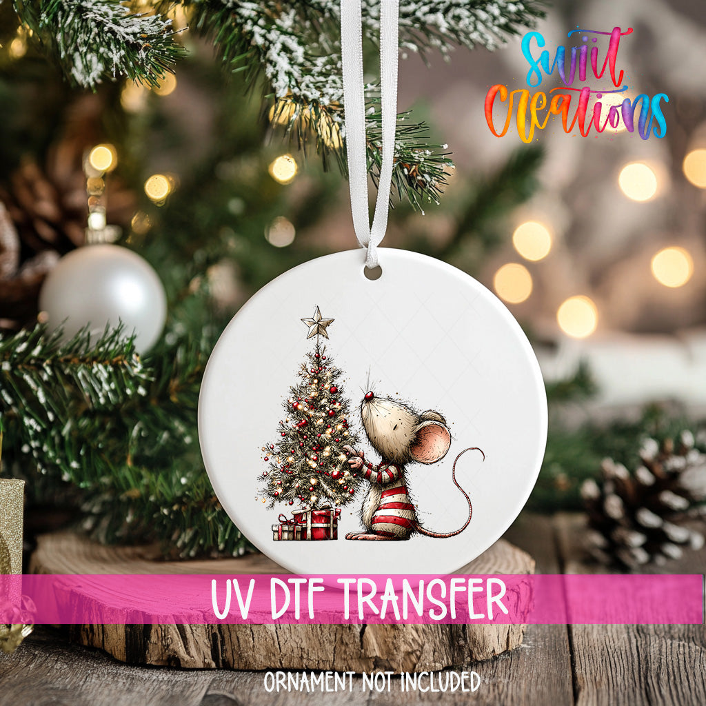 A white Christmas ornament with a mouse and a decorated Christmas tree is hanging on a tree branch.