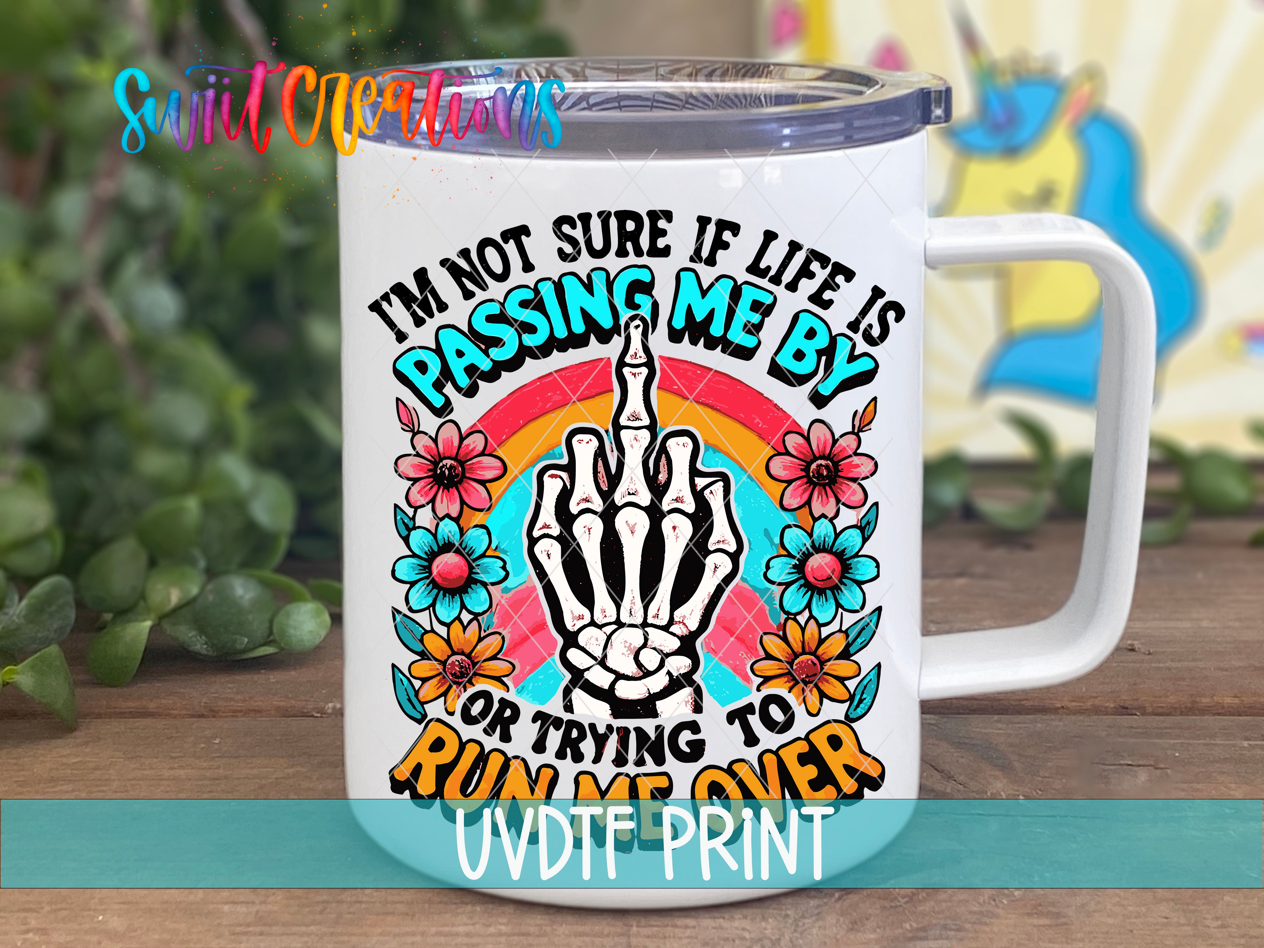 A white mug with a colorful graphic design featuring a skeleton hand, flowers, and a rainbow. The text on the mug reads "I'm not sure if life is passing me by, or trying to run me over".