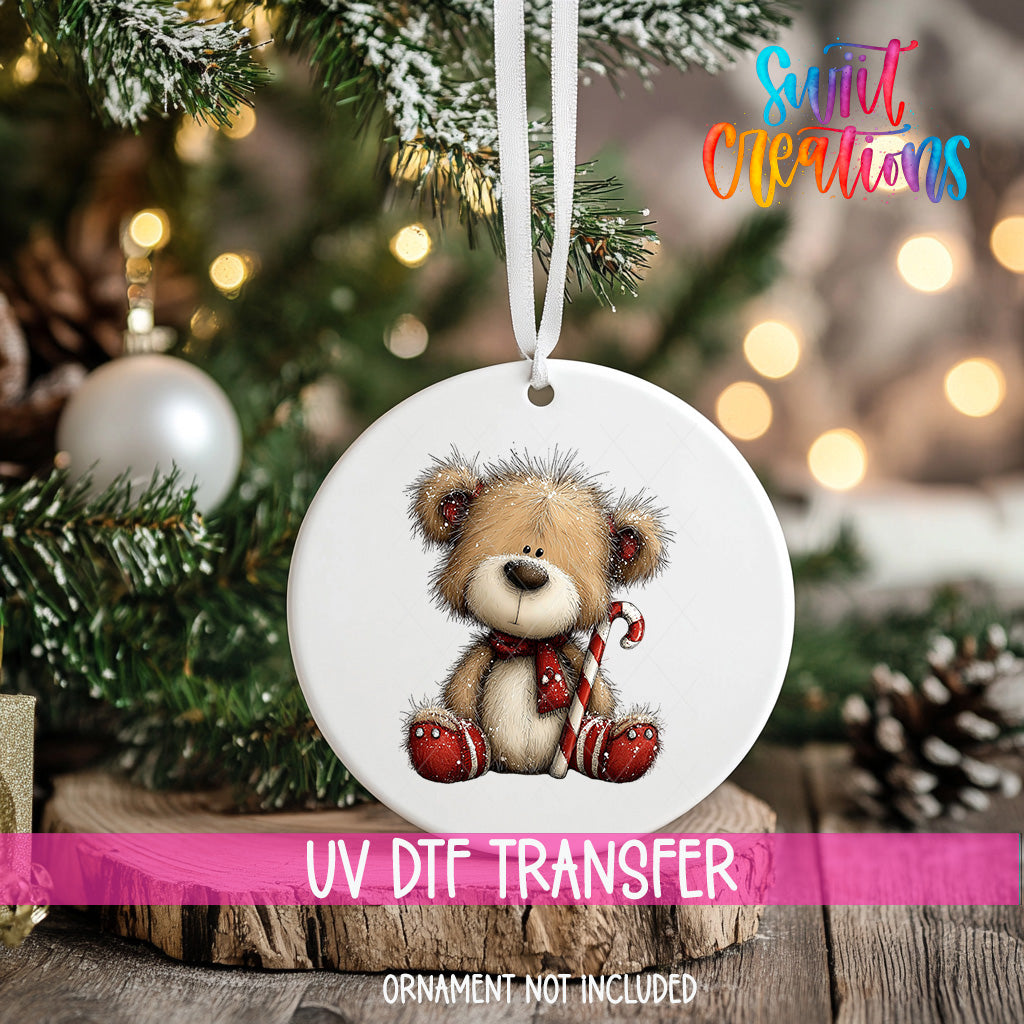 A white ornament with a teddy bear and a candy cane is hanging on a Christmas tree, with a pink ribbon around it.