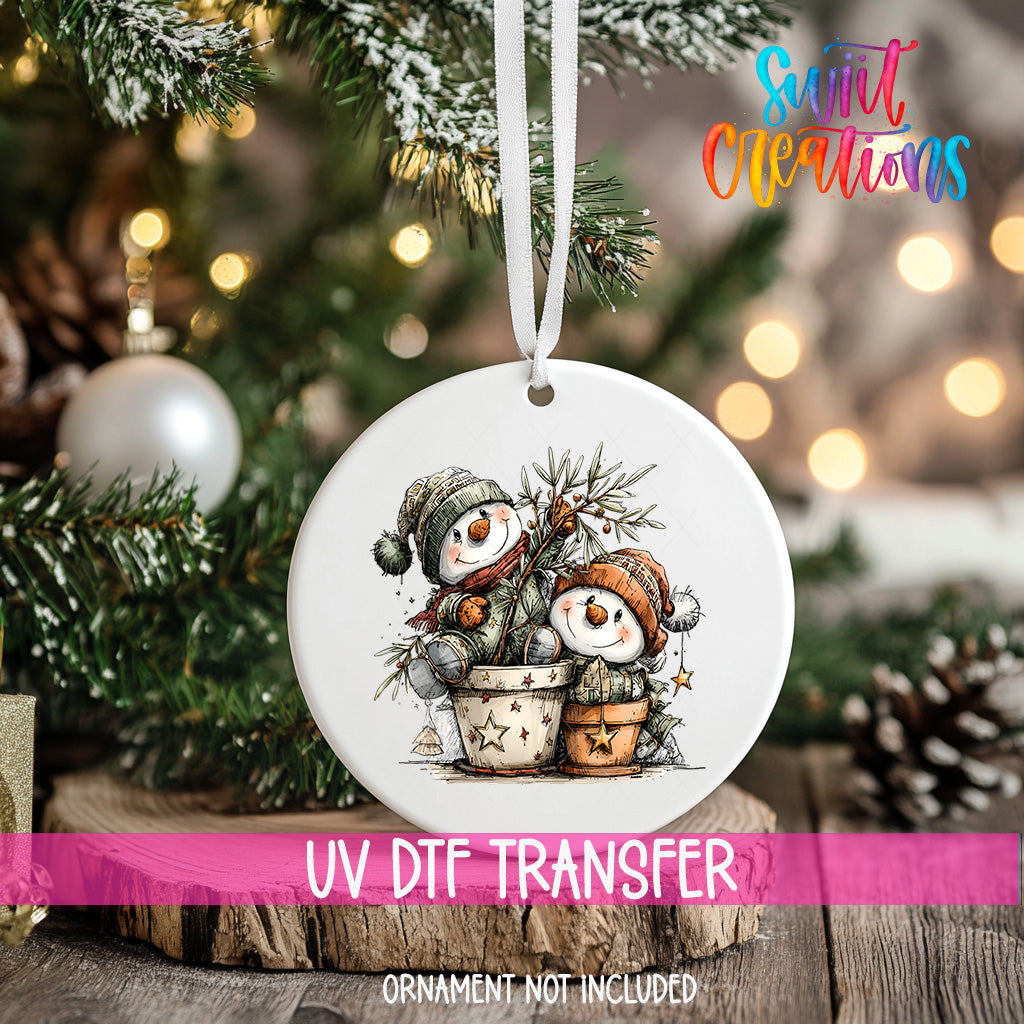 A white ornament with a snowman and pine tree design hangs on a Christmas tree, with a pink ribbon around it.