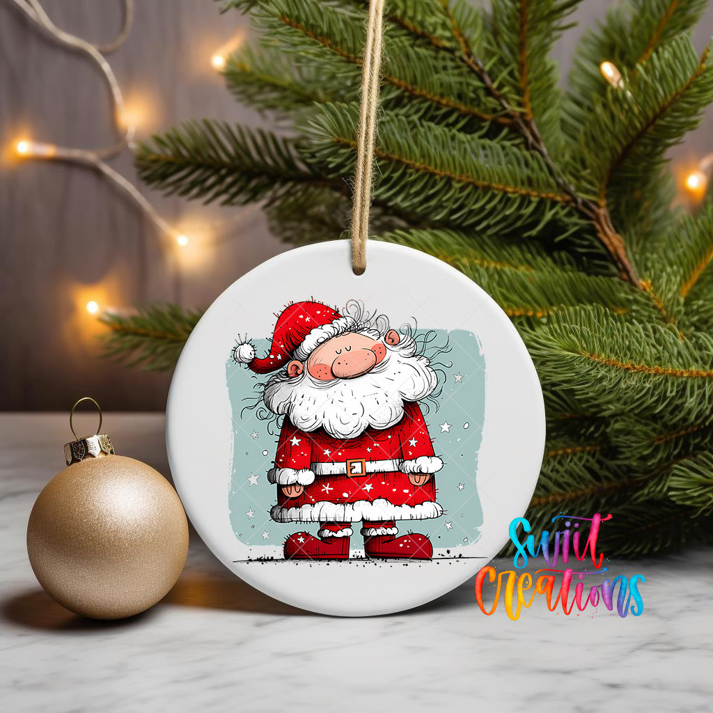 A white ceramic ornament featuring a cartoon illustration of Santa Claus wearing a red suit and hat, with snowflakes falling around him, is displayed on a table next to a gold ornament and a Christmas tree.