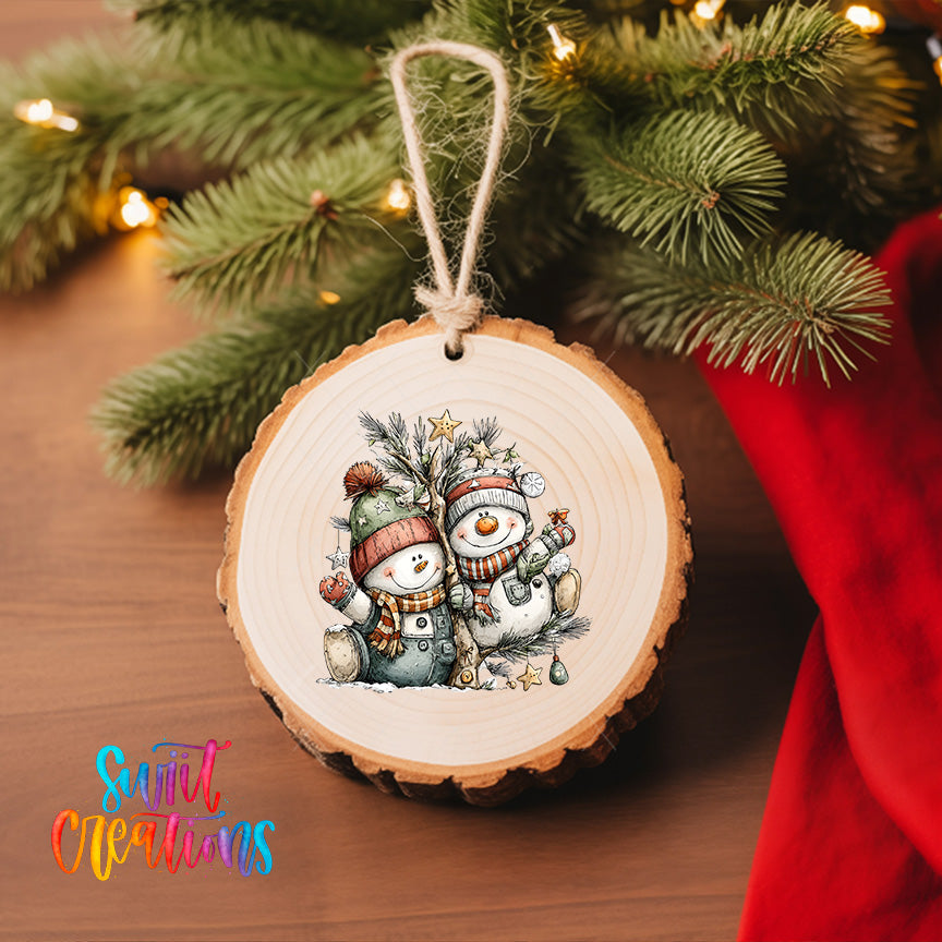 A wooden ornament featuring two snowmen is hanging on a Christmas tree, surrounded by pine branches and twinkling lights.