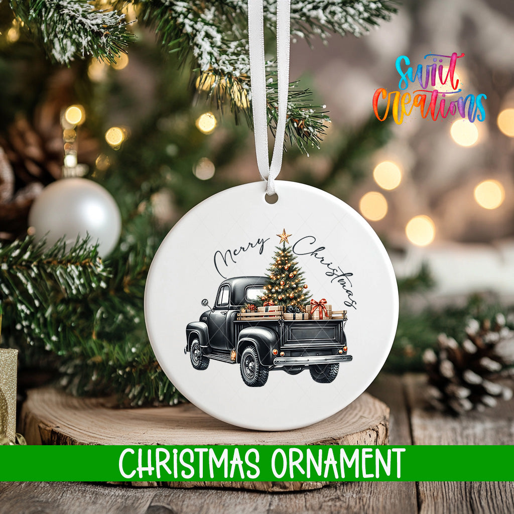 A white Christmas ornament with a black vintage truck carrying a decorated Christmas tree is hanging on a tree.