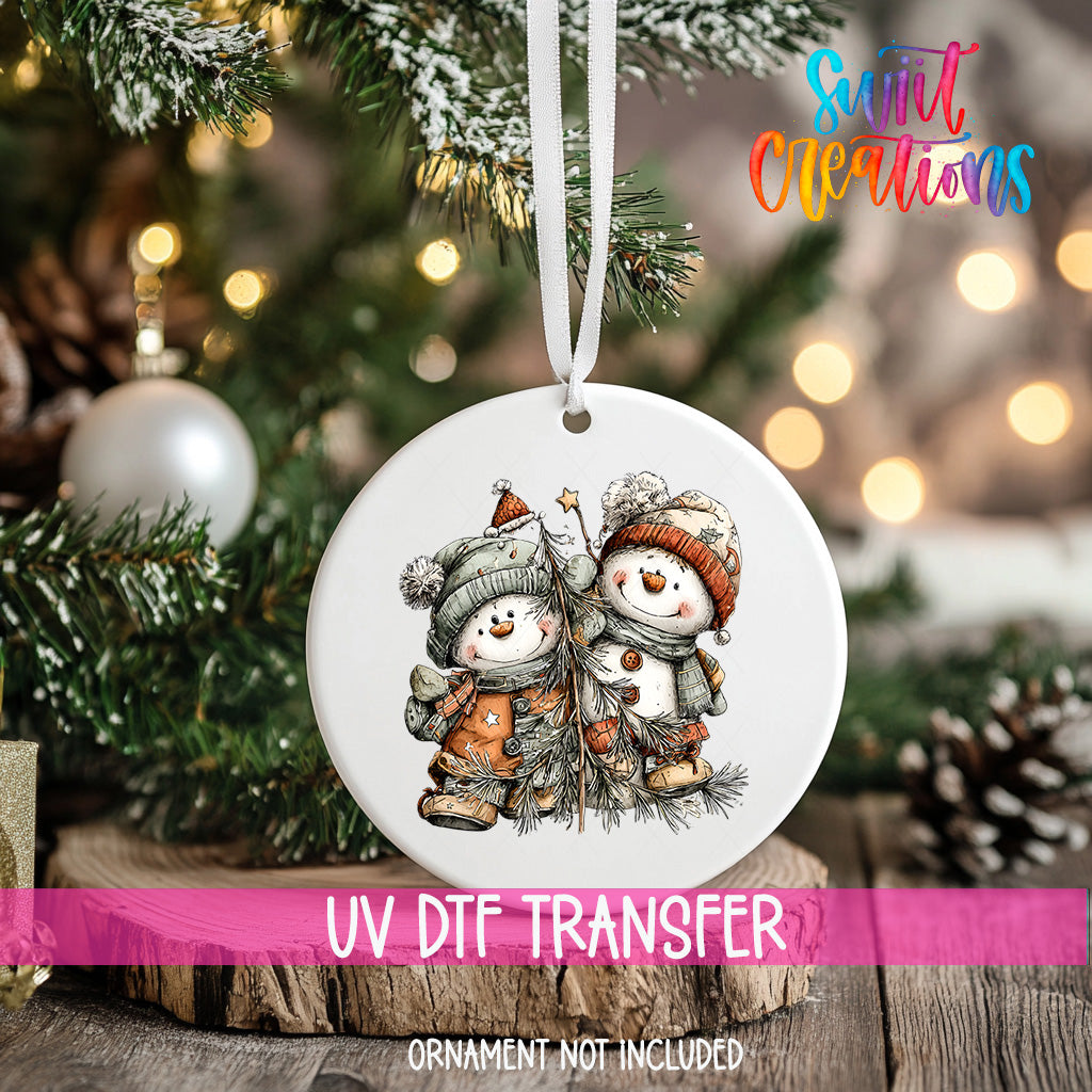 A white ornament with a snowman and pine tree design hangs on a Christmas tree, with a pink ribbon around it.