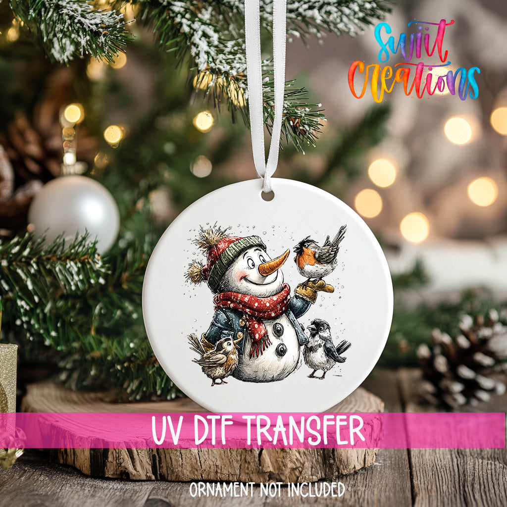 A white ornament with a snowman and birds on a Christmas tree, with a pink ribbon around it.