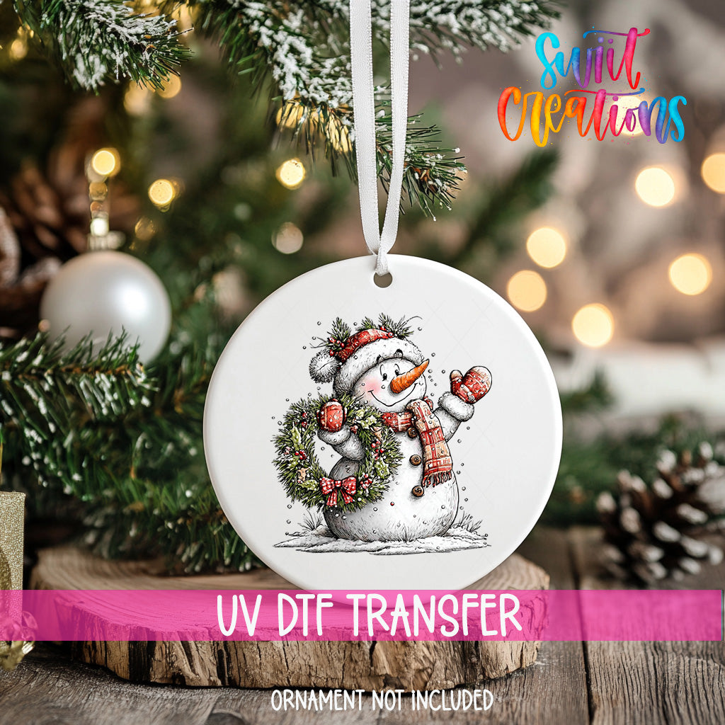 A white ornament with a snowman design hangs on a Christmas tree, surrounded by pine branches and twinkling lights.