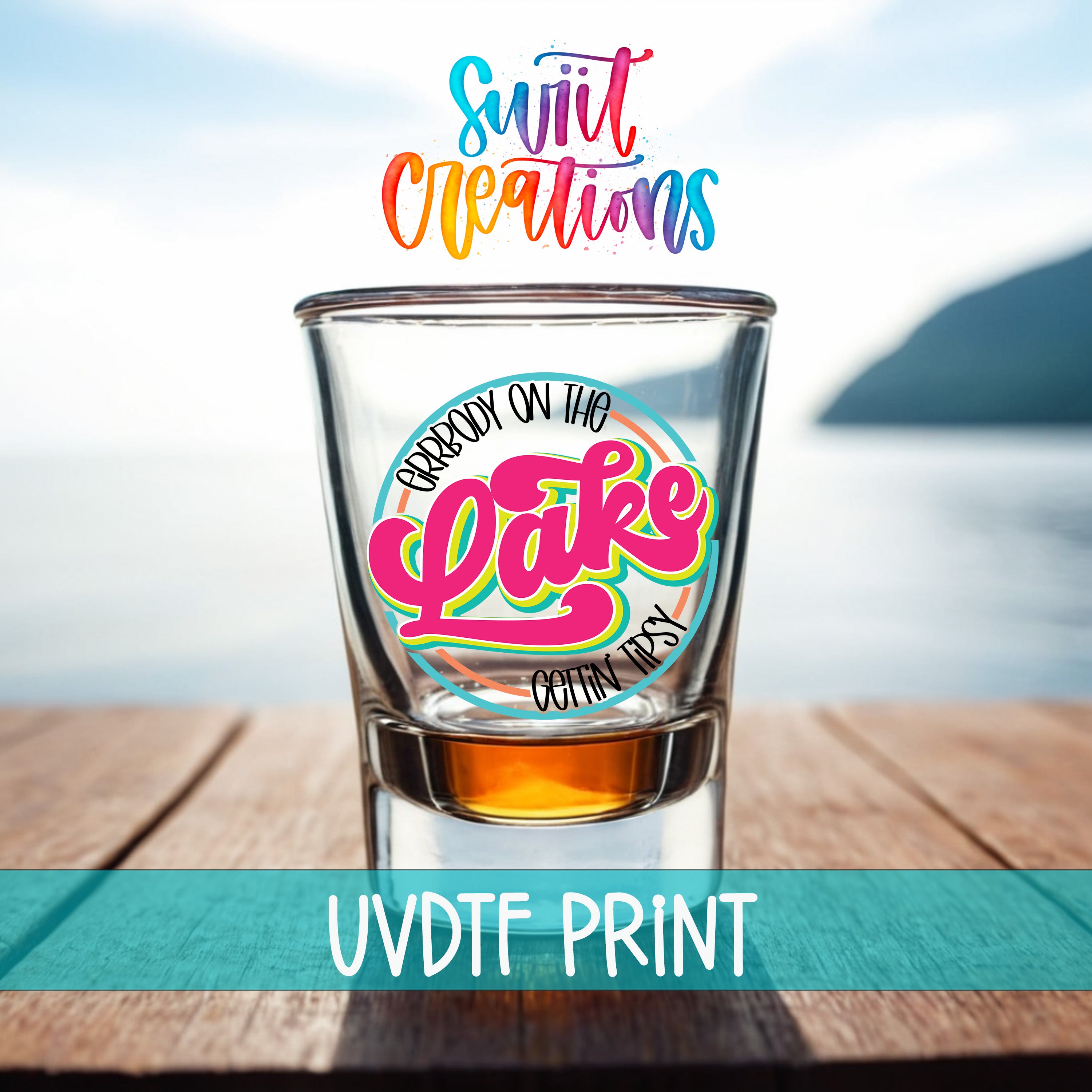 A shot glass with a colorful design on it, placed on a wooden surface with a body of water in the background.