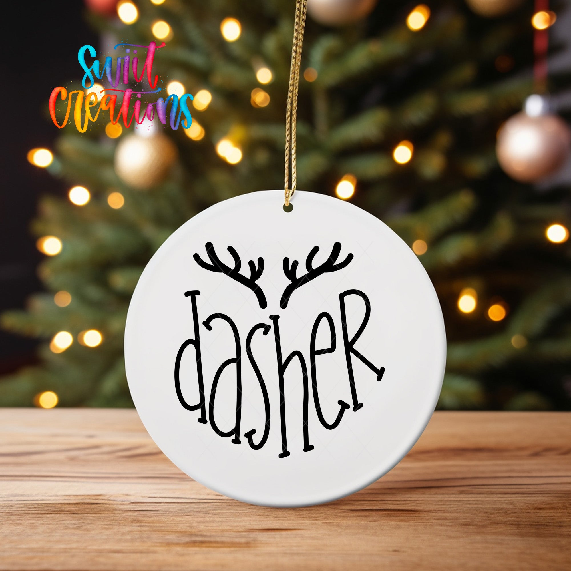 A white ornament with the word "dasher" written on it, hanging from a string in front of a blurred Christmas tree with colorful lights.