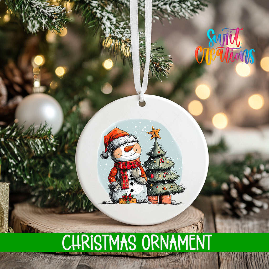 A white Christmas ornament featuring a snowman and a Christmas tree, hanging on a tree branch with a blurred background of lights.