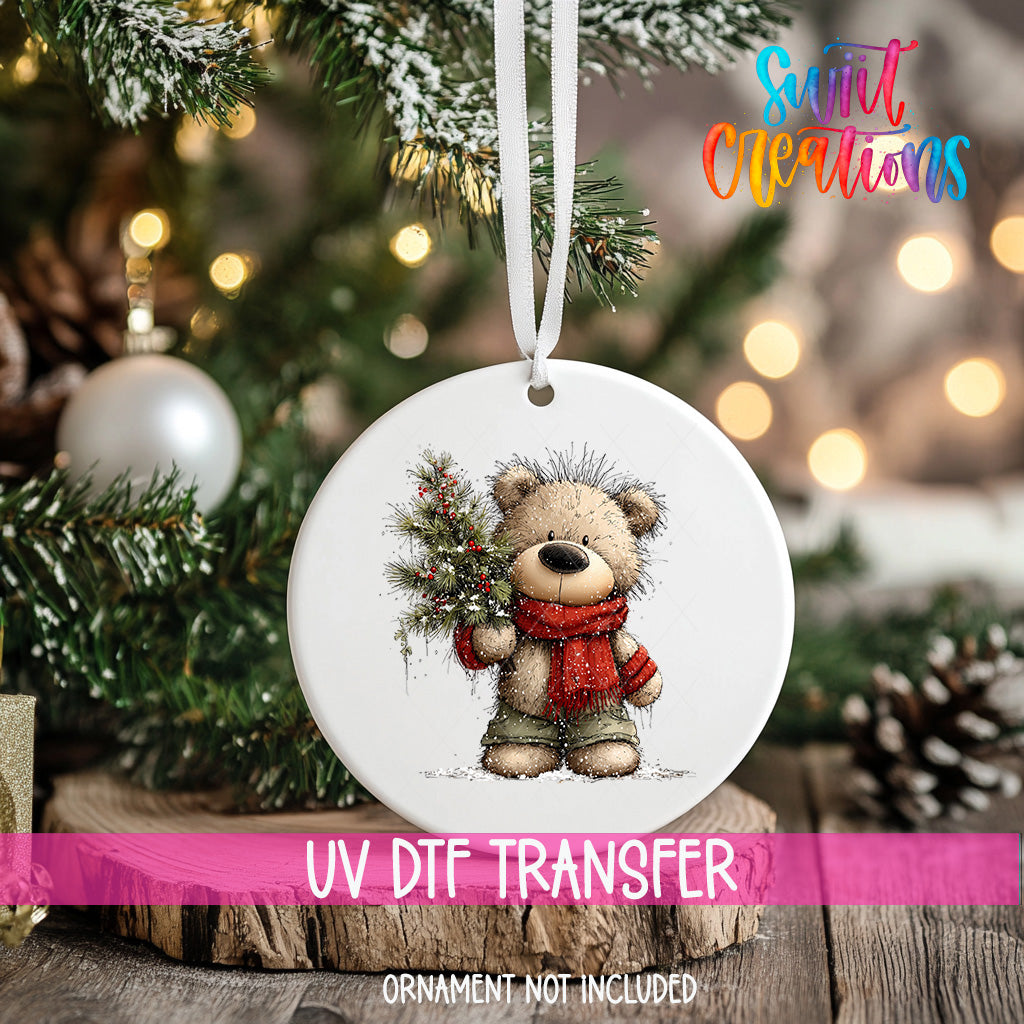 A white ornament with a teddy bear and Christmas tree is hanging on a tree, with a pink ribbon around it.