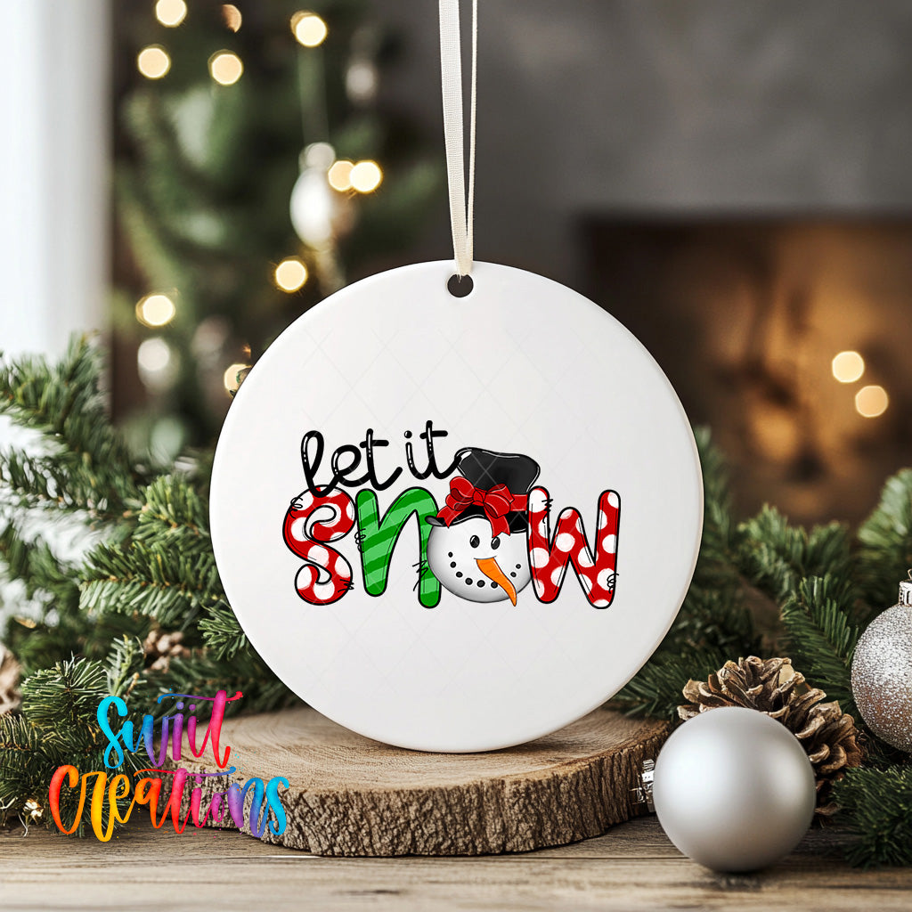 A white ceramic ornament with the text "Let it snow" and a snowman graphic, hanging on a string. The ornament is placed on a wooden surface, surrounded by Christmas decorations such as pine cones and ornaments.