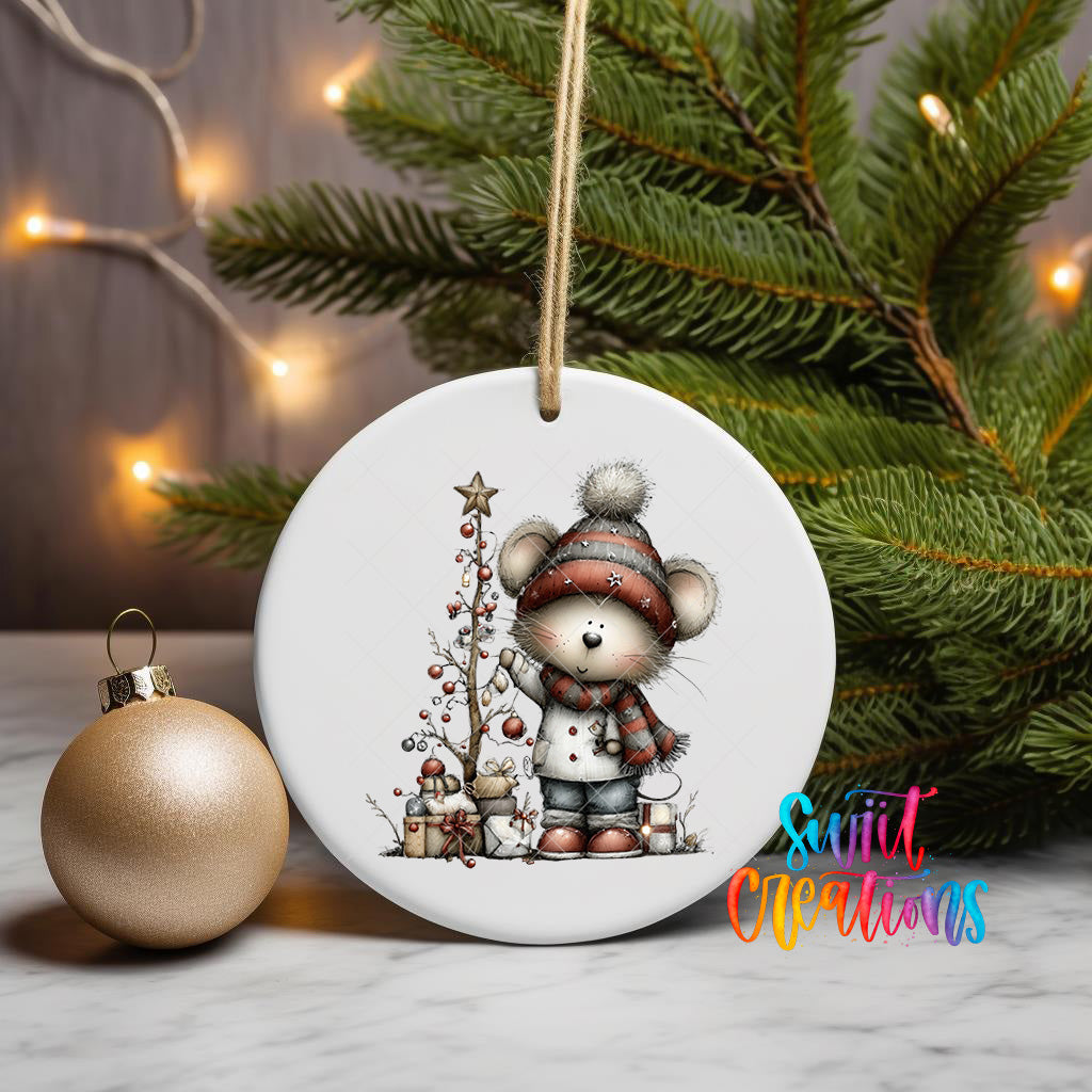 A white ceramic ornament featuring a cartoon mouse wearing a winter hat and scarf, standing next to a Christmas tree and presents, with a gold ornament and twinkling lights in the background.