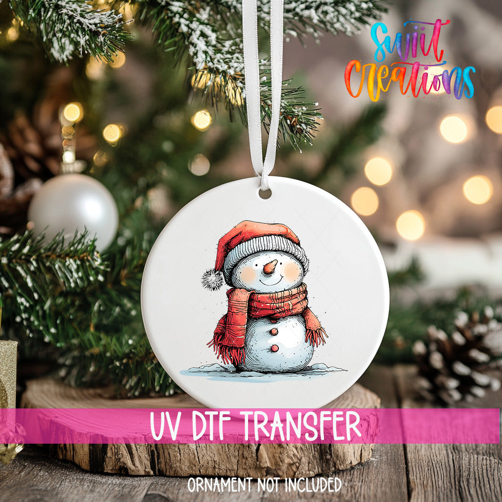 A white ornament with a snowman design hangs on a Christmas tree, surrounded by pine branches and twinkling lights.