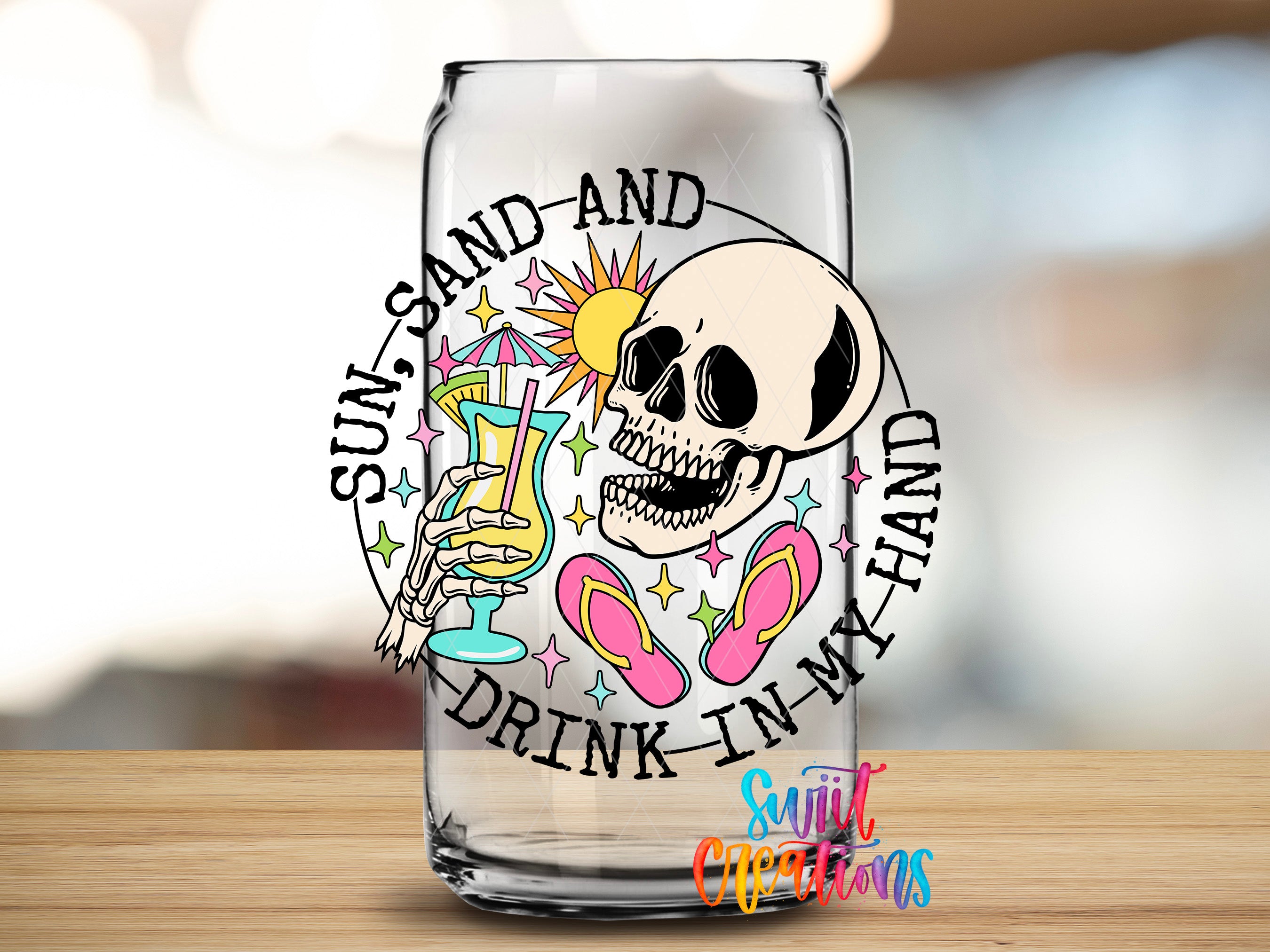 A glass with a colorful design featuring a skull, sunglasses, a beach umbrella, and a cocktail glass, along with the text "Sun, sand, and drink in my hand".