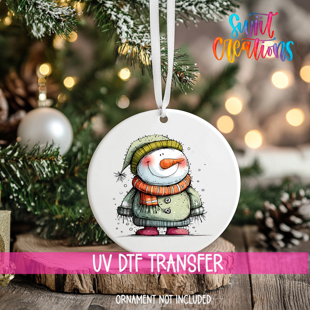 A white ornament with a snowman design hangs on a Christmas tree, surrounded by pine branches and twinkling lights.