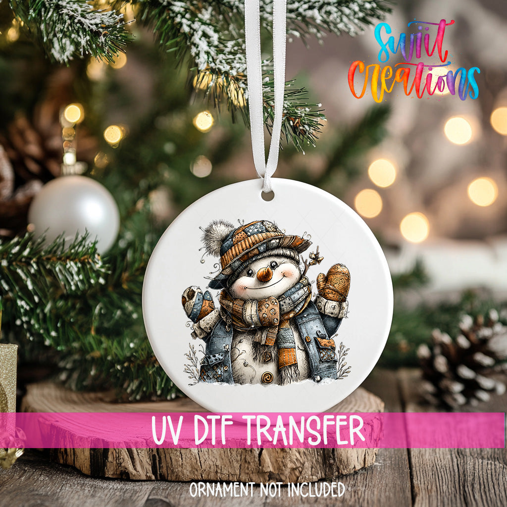 A white ornament with a snowman design hangs on a Christmas tree, surrounded by pine branches and twinkling lights.