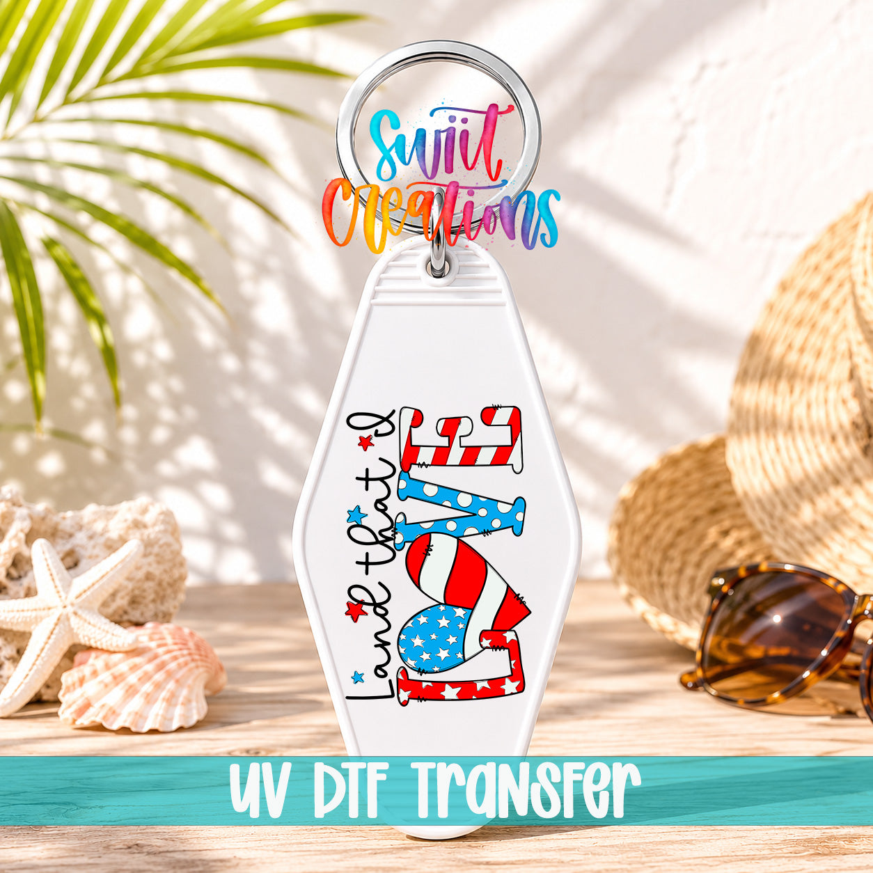 A keychain with a patriotic design, including a red, white, and blue heart shape, and the text "Land that I Love" printed on it. The keychain is placed on a wooden surface, with sunglasses and a straw hat visible in the background.