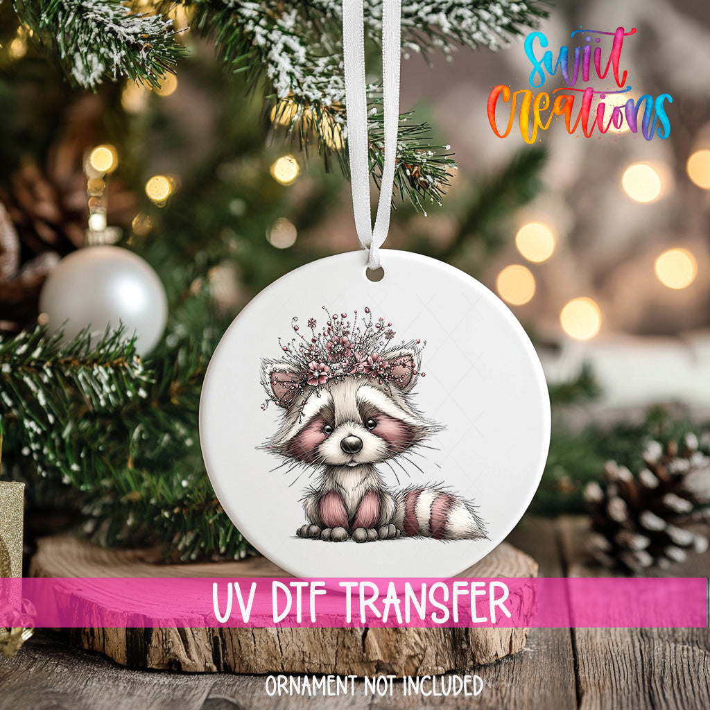 A white ornament with a cute raccoon design hangs on a Christmas tree, surrounded by pine branches and twinkling lights.