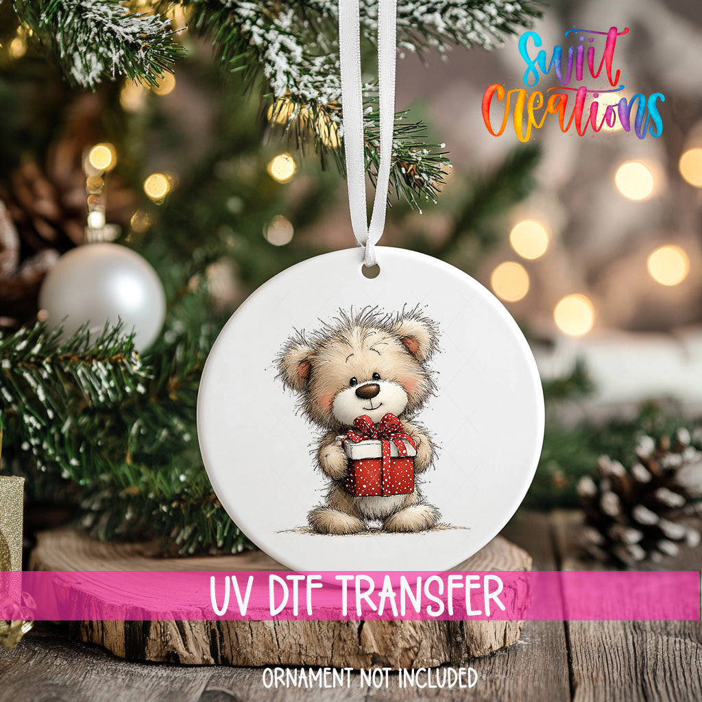 A white ornament with a teddy bear holding a gift is hanging on a Christmas tree, with a blurred background of pine branches and twinkling lights.