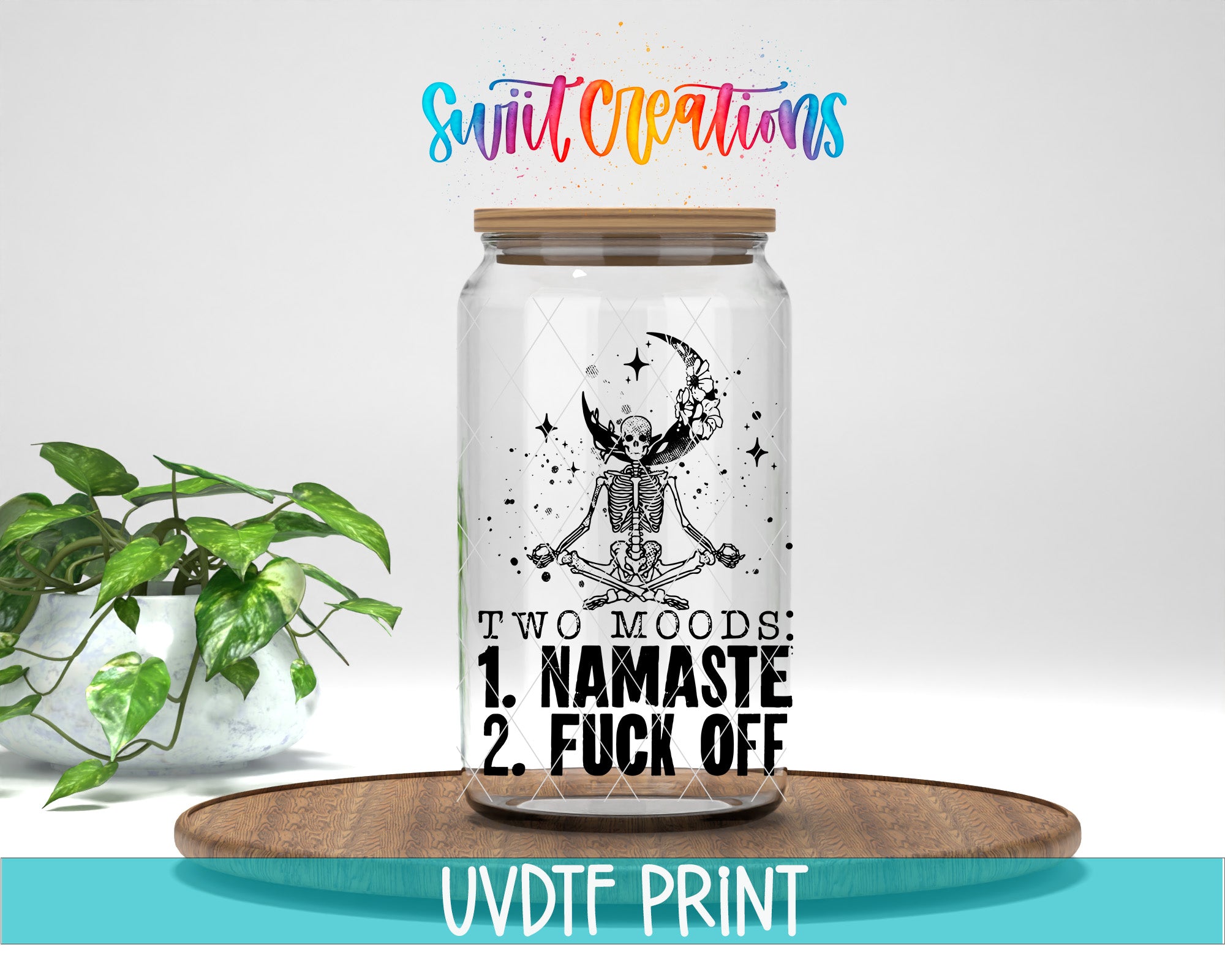 A glass jar with a skeleton and crescent moon design, along with the text "TWO MOODS 1. NAMASTE 2. FUCK OFF" printed on it. The jar is placed on a wooden tray, and there is a potted plant in the background.