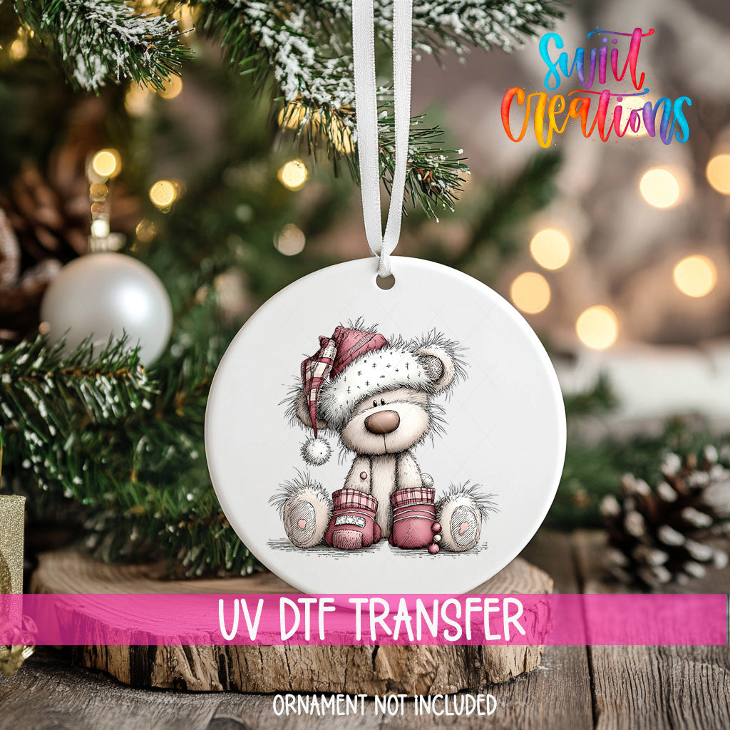 A white ceramic ornament with a teddy bear wearing a red hat and boots, sitting on a wooden surface, with a blurred background of a Christmas tree and twinkling lights.