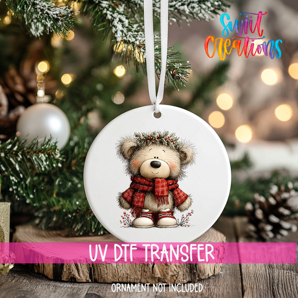 A white ornament with a teddy bear wearing a red scarf and a crown, hanging on a Christmas tree.