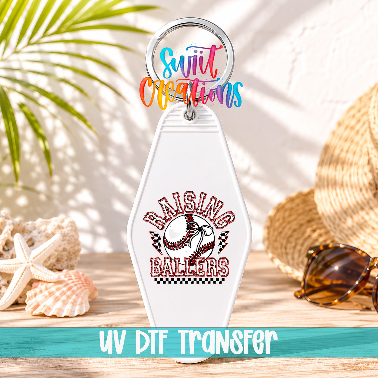 A keychain with a baseball design and the text "Raising Ballers" printed on it, along with a colorful ring and a straw hat. The keychain is placed on a wooden surface, with sunglasses and a palm frond visible in the background.