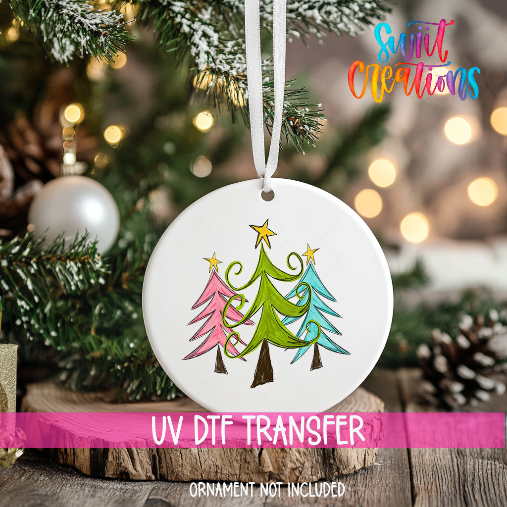 A white Christmas ornament with a colorful design of three trees hangs on a tree branch, with a blurred background of lights and greenery.