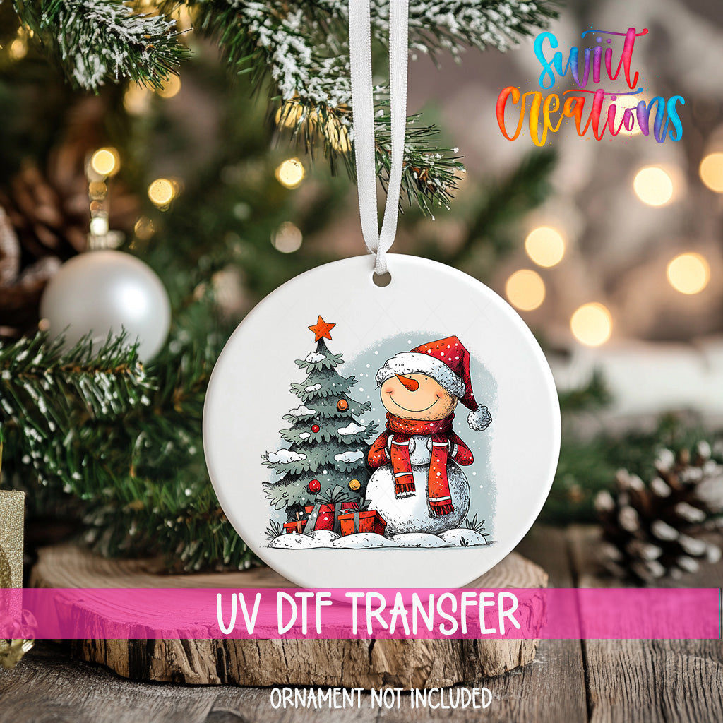 A white Christmas ornament with a snowman and a Christmas tree, hanging on a tree branch.