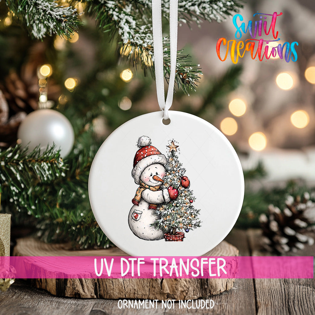 A white Christmas ornament with a snowman and a Christmas tree is hanging on a tree, with a pink ribbon around it.