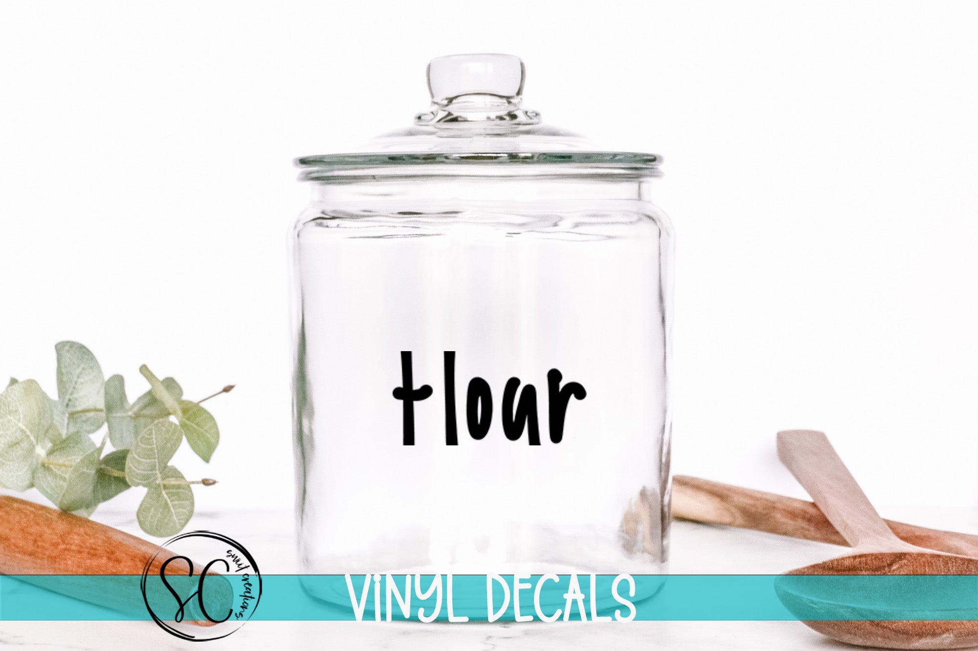 A clear glass jar with a black label that says "flour" is placed on a white surface, surrounded by various kitchen utensils and decorative items.
