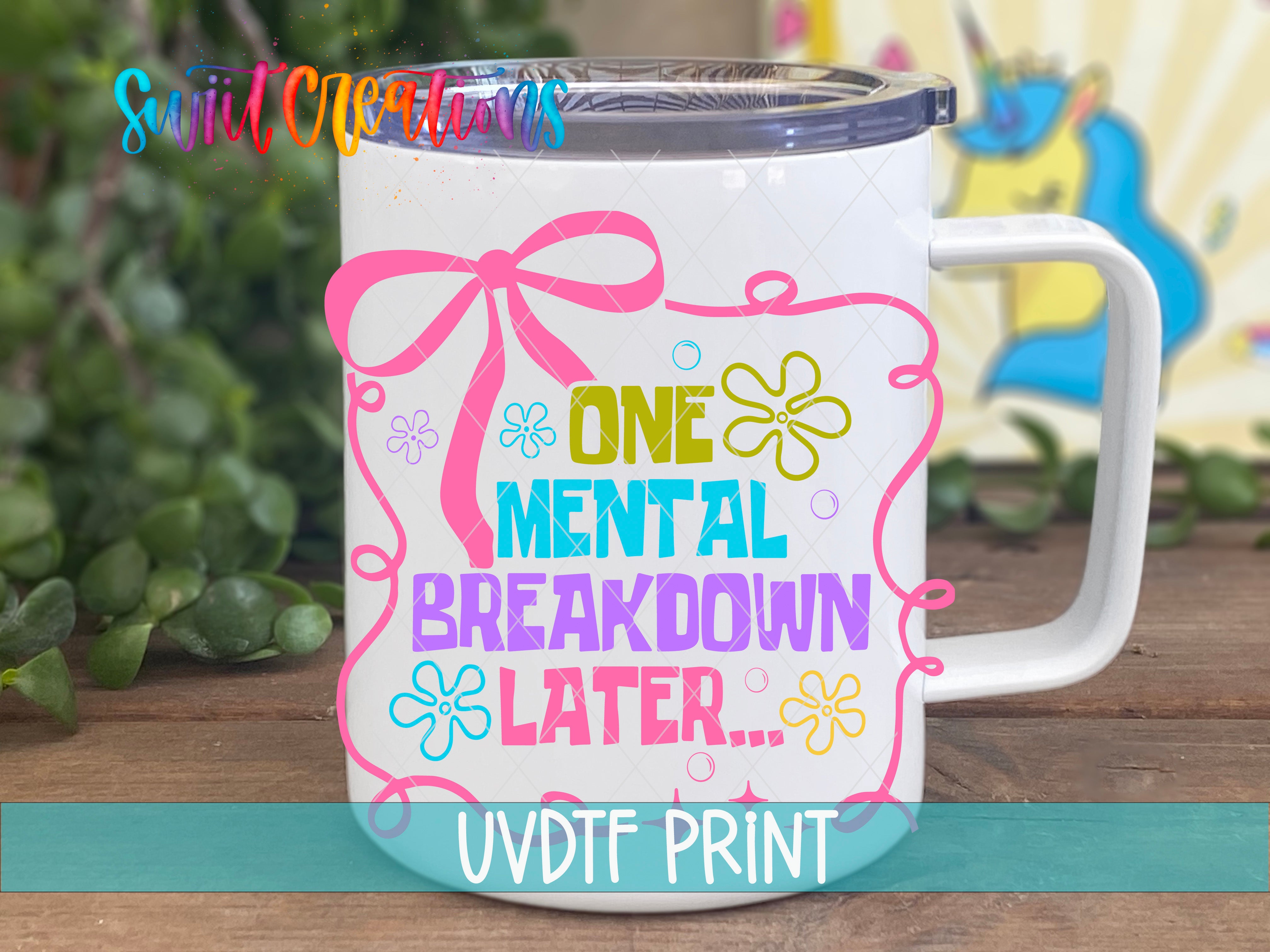 A white mug with a colorful graphic design featuring text and flowers, placed on a wooden surface with greenery in the background.