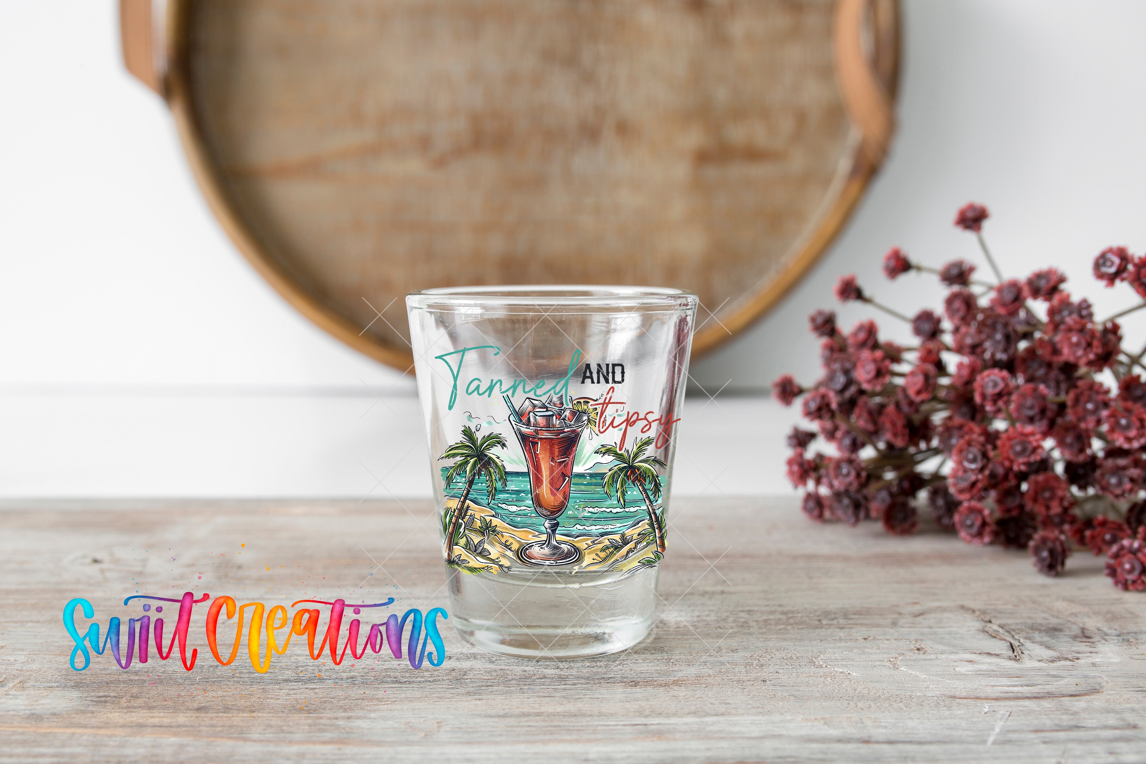A shot glass with a tropical beach scene and palm trees printed on it, placed on a wooden surface next to a bunch of red flowers.
