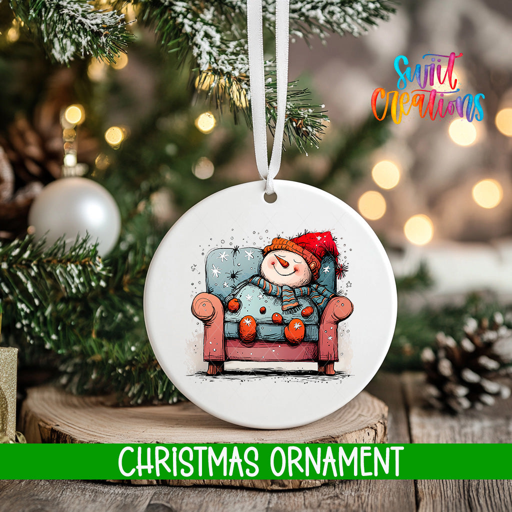 A white Christmas ornament with a snowman sitting in a chair, surrounded by snow and twinkling lights, is displayed on a tree branch.