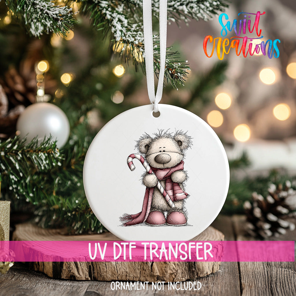 A white ornament with a teddy bear and a candy cane is hanging on a Christmas tree, with a pink ribbon around it.