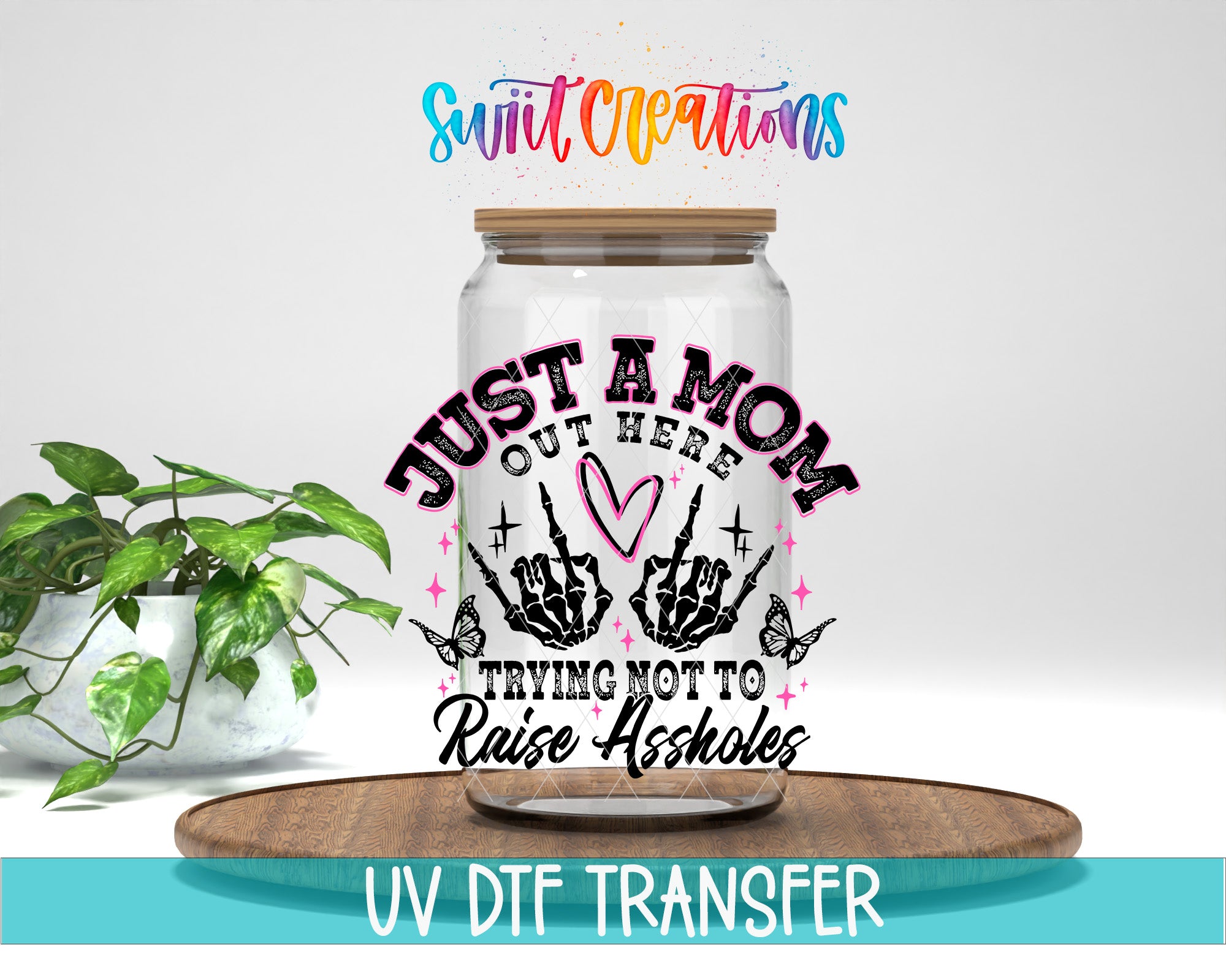 A glass jar with a printed design on it, placed on a wooden surface. The jar has a lid and is surrounded by a plant. The text on the jar reads "Just a Mom, out here trying not to raise assholes".