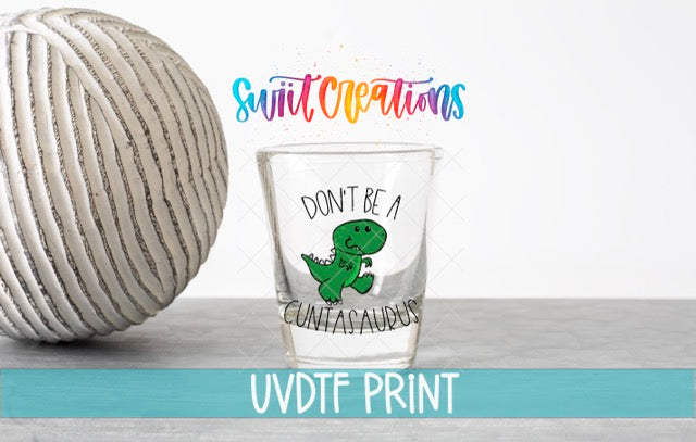 a glass with a dinosaur design and text, placed next to a white ball on a gray surface.