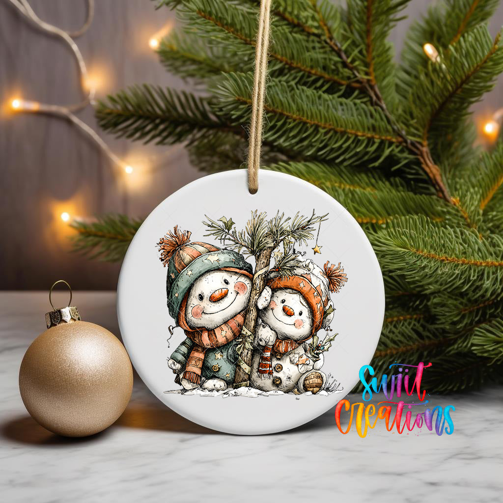 A white ceramic ornament featuring two cheerful snowmen, one wearing a green scarf and the other a red scarf, sitting together under a tree. The ornament is hanging from a string, and there is a gold ornament and a Christmas tree in the background.