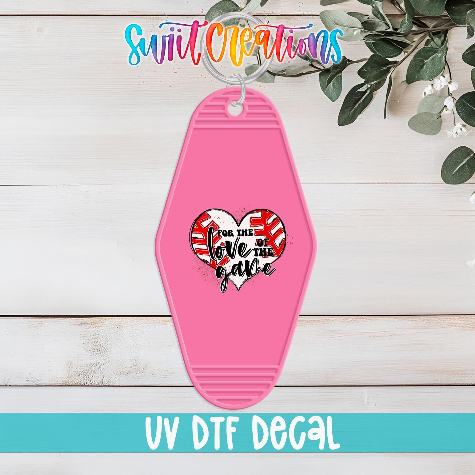 A pink keychain with a heart-shaped design and text on it, placed on a wooden surface with some greenery in the background.