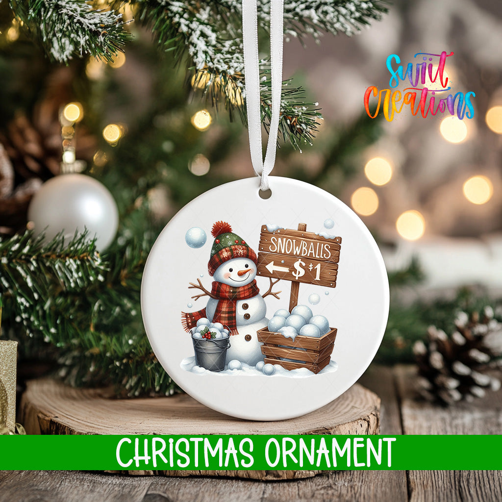 A white Christmas ornament featuring a snowman holding a sign that says "Snowballs $1" and a bucket of snowballs, hanging on a tree branch with a blurred background of Christmas lights.