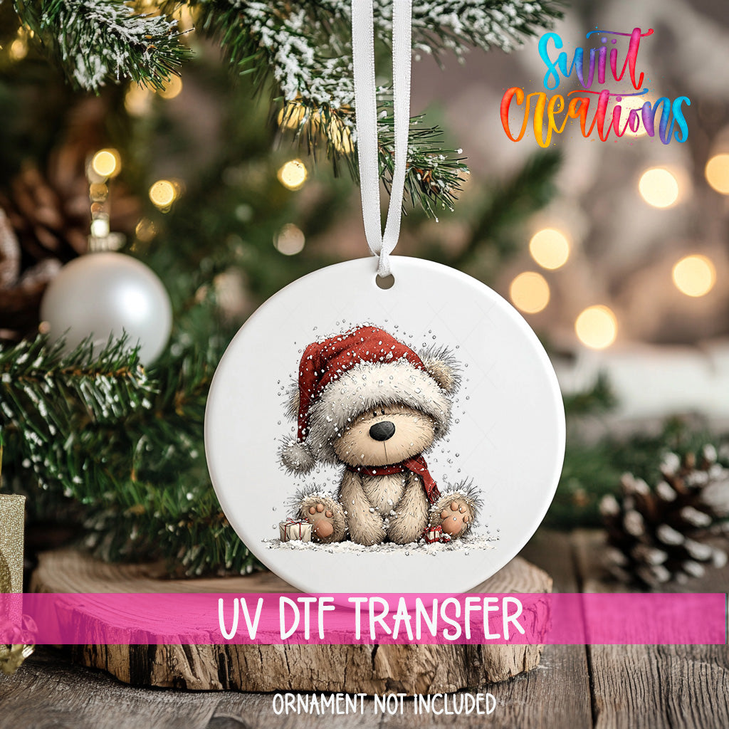 A white Christmas ornament with a teddy bear wearing a Santa hat, hanging on a tree branch with a blurred background of lights.