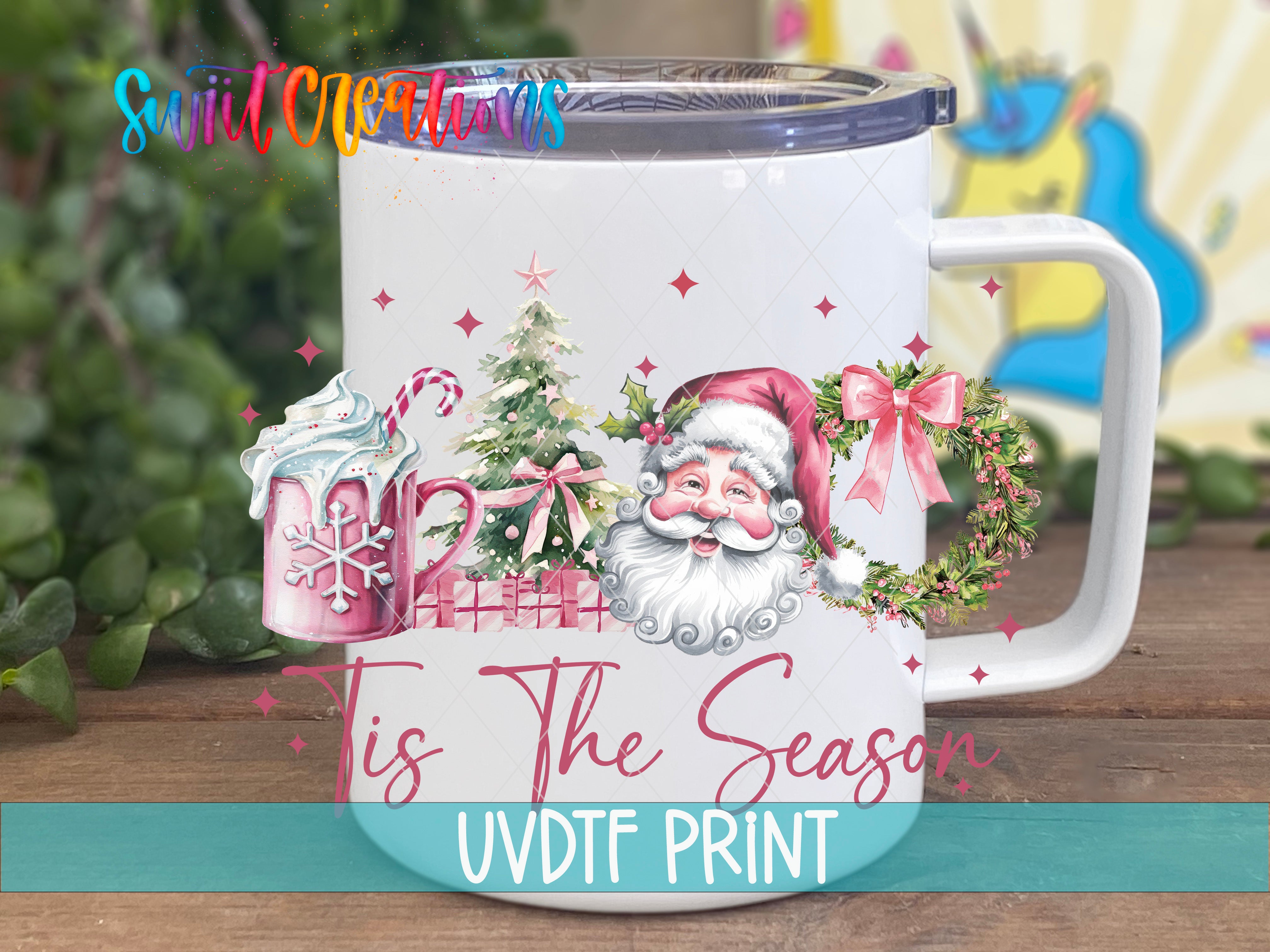 A white mug with a Christmas-themed design, including a Santa Claus face, a snowflake, and a wreath. The mug is placed on a wooden surface, and there is a blurred background that appears to be a garden or outdoor setting.