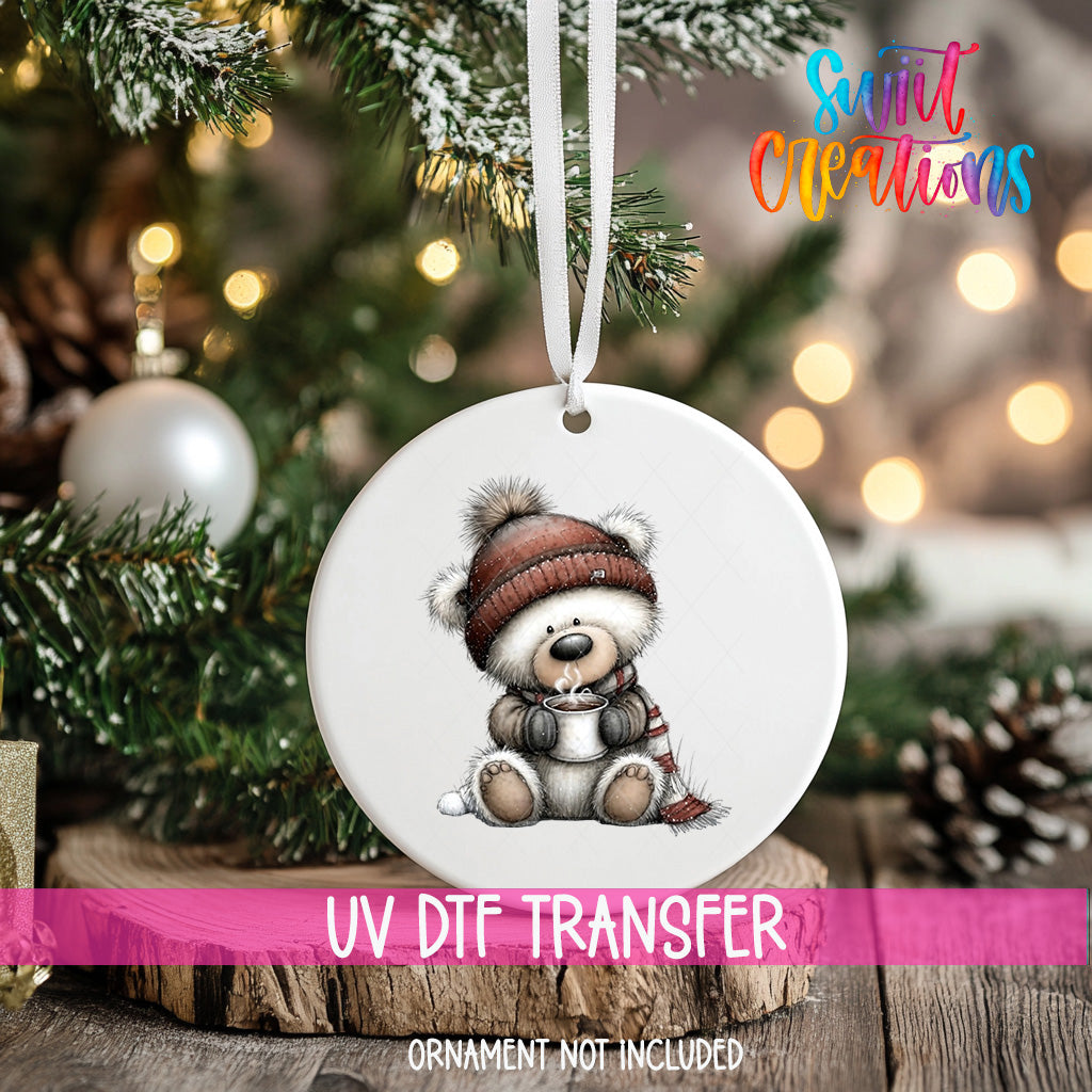 A white ornament with a teddy bear wearing a hat and scarf is hanging on a Christmas tree.