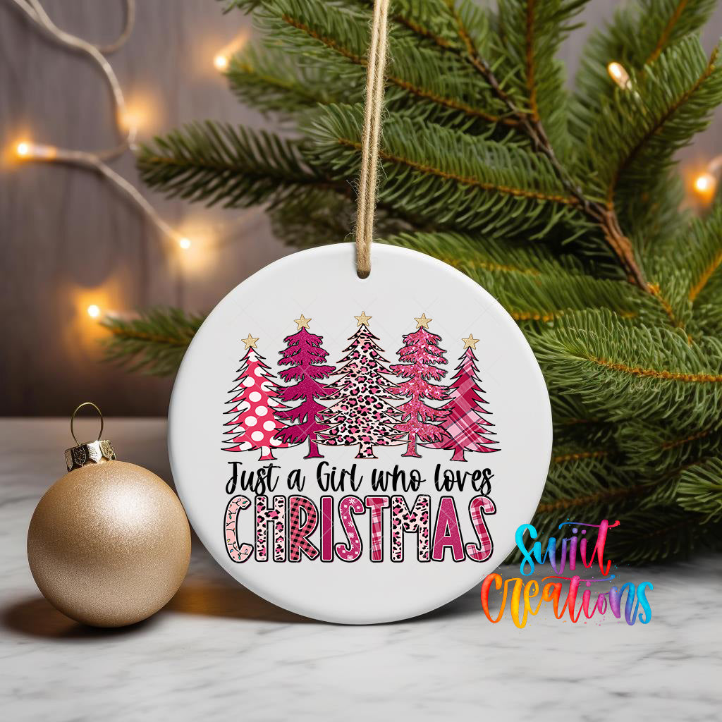 A white Christmas ornament with a design of pink and white trees, along with the text "Just a girl who loves Christmas" is displayed on a table, with a gold ornament and a pine tree in the background.