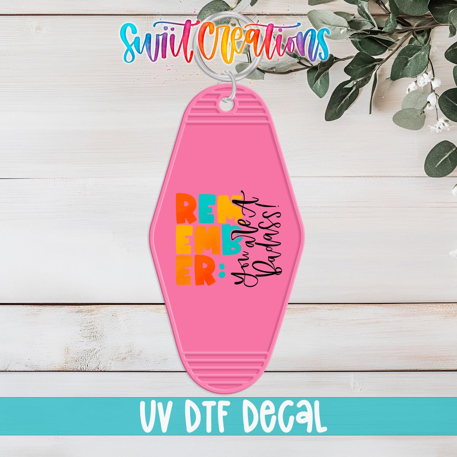 A pink keychain with the text "REMEMBER" in large letters, along with a smaller text that reads "You're all bad" in a stylized font. The keychain is displayed on a wooden surface, with some greenery visible in the background.