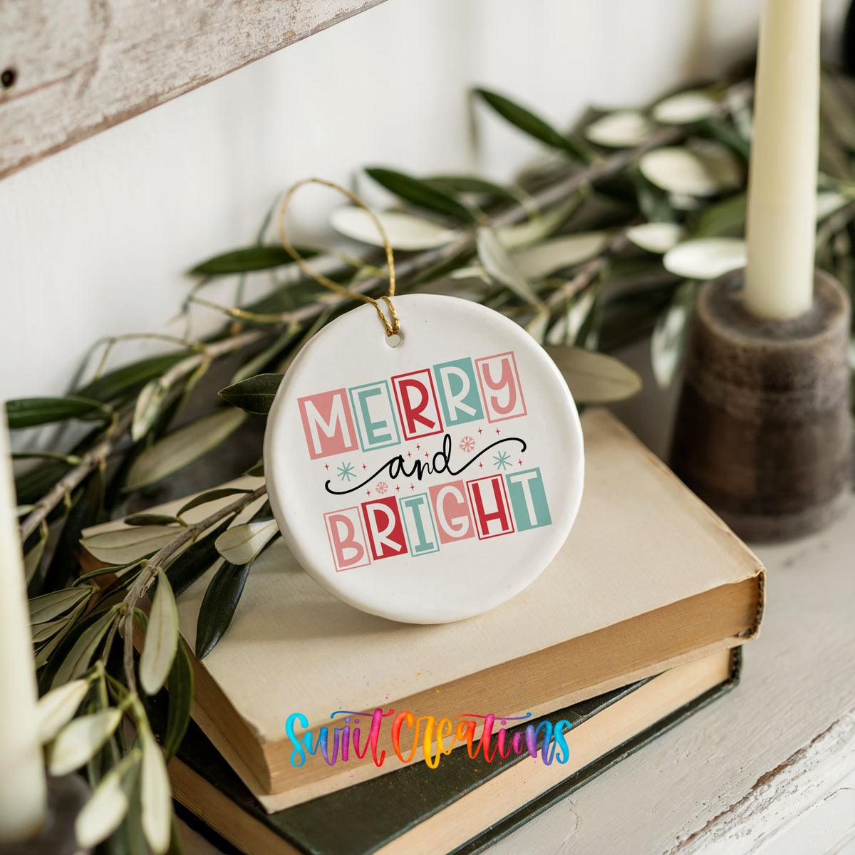 Round ceramic Christmas ornament featuring pastel block-letter Merry and Bright holiday typography, styled on books with holiday greenery